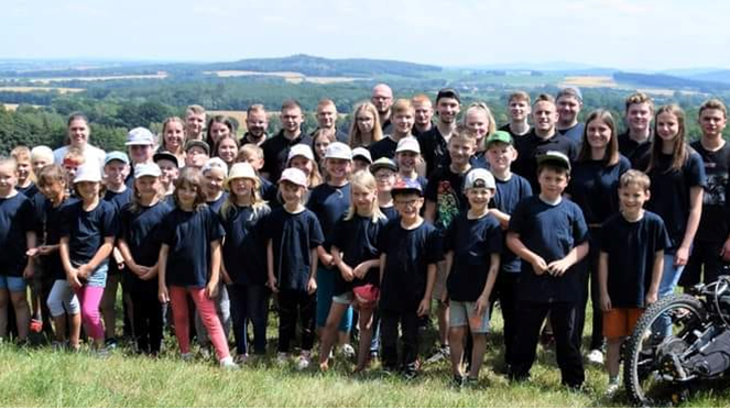 ausflugEine große Gruppe von Kindern und Erwachsenen steht draußen auf einer Wiese, im Hintergrund ist eine hügelige Landschaft zu sehen. Viele tragen schwarze T-Shirts, einige sitzen, andere stehen.